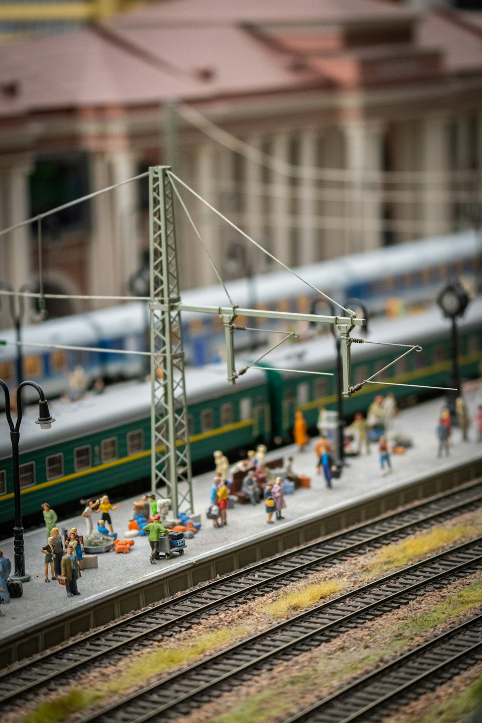 Kleine Figuren warten an einer Modellbahnanlage auf einen grünen Personenzug.